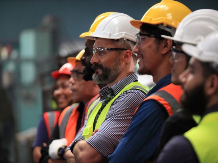 A line of smiling professionals in hard hats watching new equipment being tested in a warehouse.