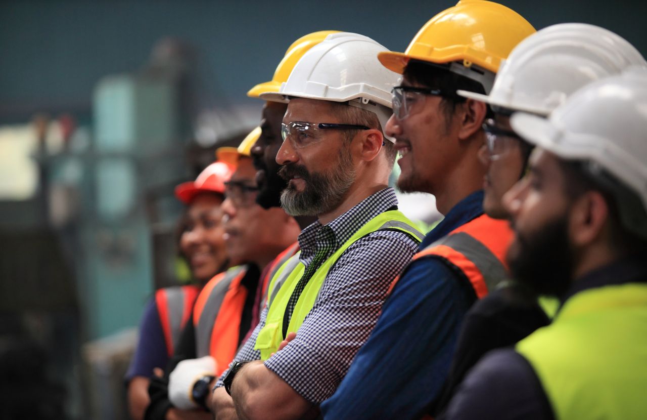 A line of smiling professionals in hard hats watching new equipment being tested in a warehouse.