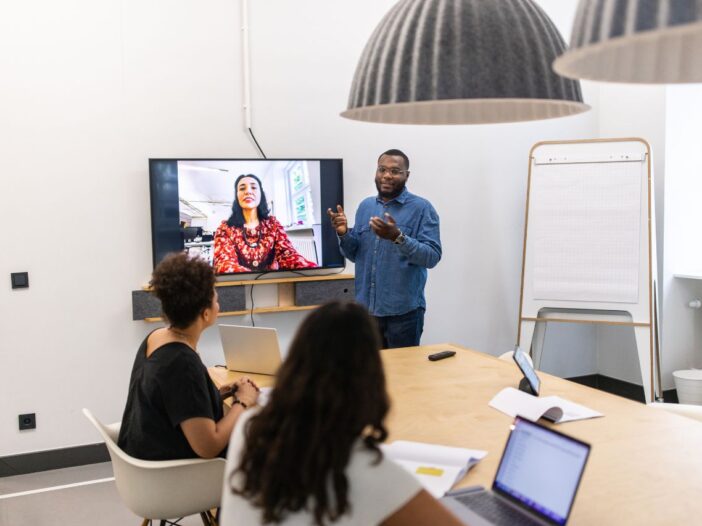 image of man presenting to three women, one of which who is working remotely