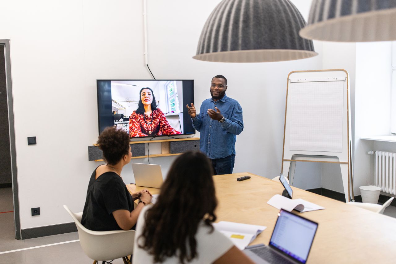 image of man presenting to three women, one of which who is working remotely