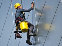 Workman in hard hat and safety harness working at height cleaning smart office block exterior.