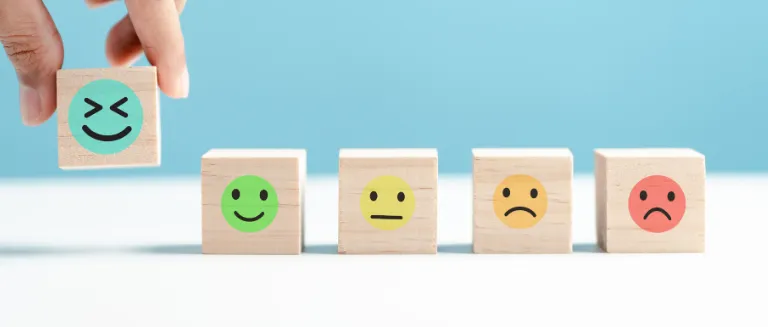 Hand lining up small wooden blocks displaying happy, sad, neutral and laughing face emojis.