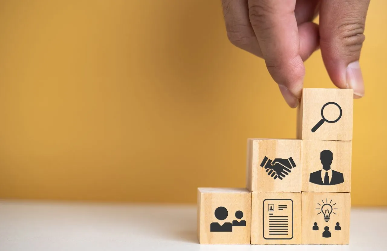 Hand stacking wooden blocks in a stair formation. The blocks display a magnifying glass, hand shake, person, notepad, and lightbulb above people's heads.