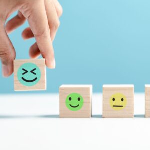 Three wooden cubes showing different facial expressions, laughing, happy, neutral. Hand picks up laughing cube.