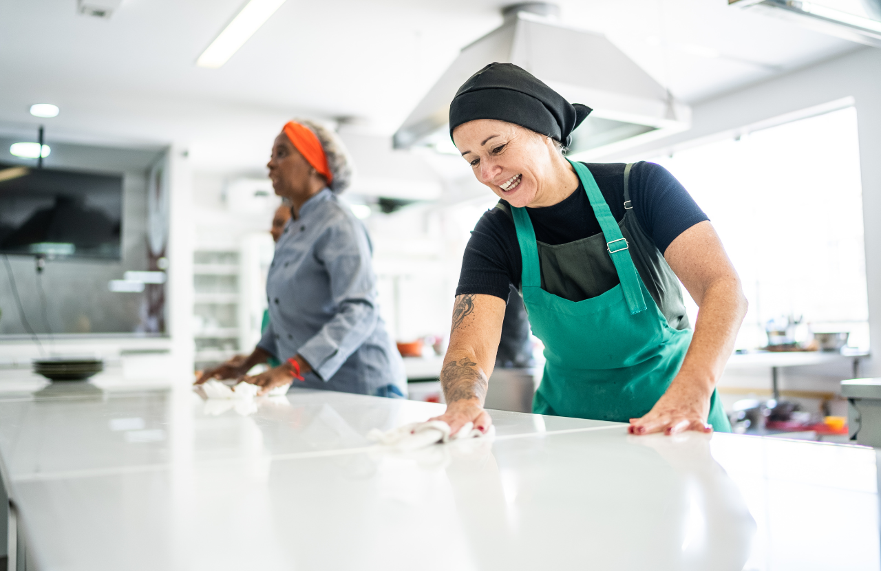 Two women in a commercial kitchen cleaning a surface to illustrate 'clean as you go'