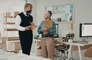 Two people talking in an office, standing up. One holding a clipboard carrying out a workplace stress risk assessment