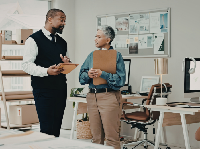 Two people talking in an office, standing up. One holding a clipboard carrying out a workplace stress risk assessment