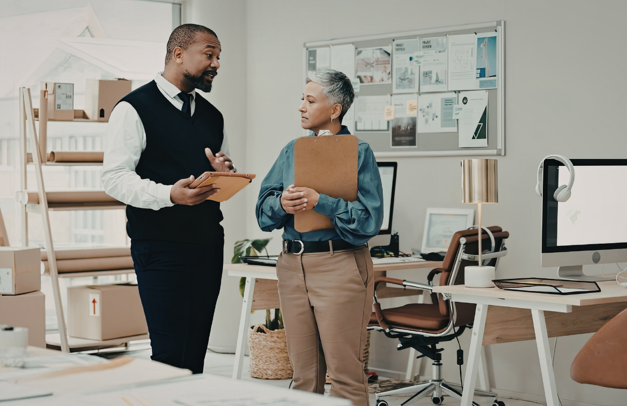 Two people talking in an office, standing up. One holding a clipboard carrying out a workplace stress risk assessment