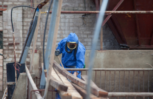 Man in full PPE removing asbestos from roof space to illustrate asbestos register.