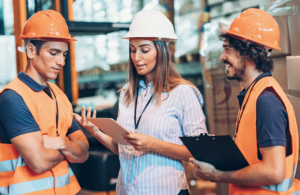Two men and a women in PPE in a warehouse smiling and talking looking at an ipad to represent health and safety report
