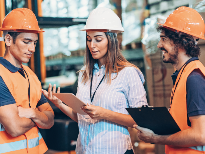 Two men and a women in PPE in a warehouse smiling and talking looking at an ipad to represent health and safety report